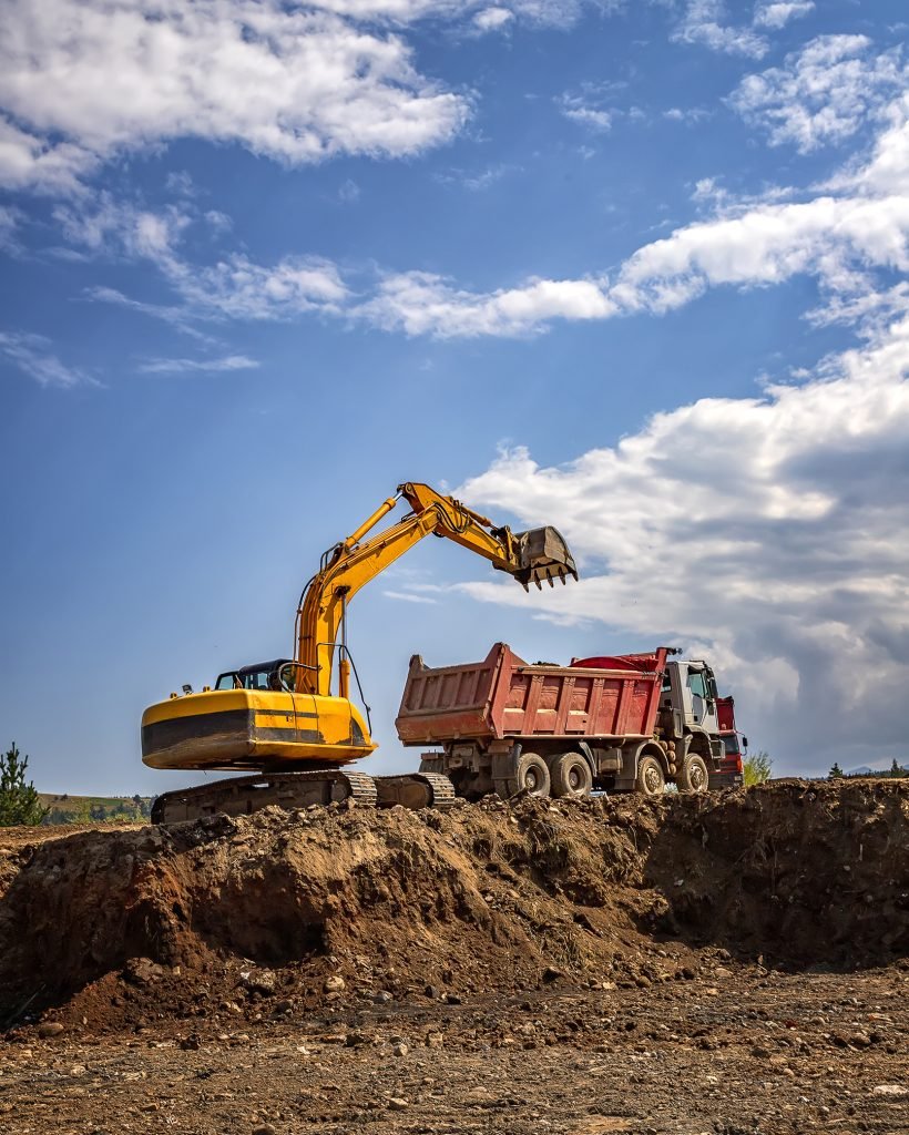 Yellow excavator and empty dump truck working at the construction site. Vertical view